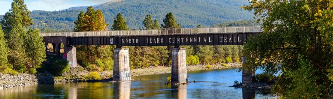 The North Idaho Centennial Trail bridge crossing the Spokane River on concrete piers, its name lettered in white on the black steel span.