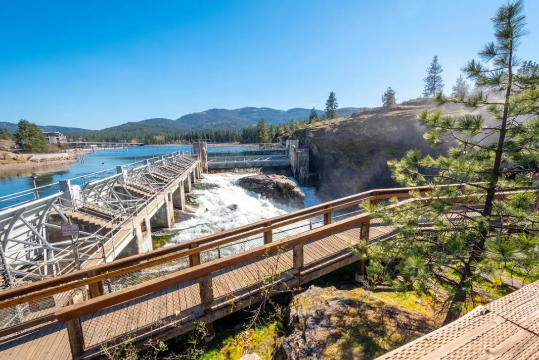 The North Channel Dam at Post Falls spilling Spokane River water, seen from the Falls Park boardwalk on a clear day.