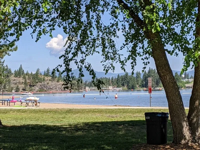 The swimming beach at Q'emiln Park on the Spokane River in Post Falls, with the river and distant dam seen through shade trees.
